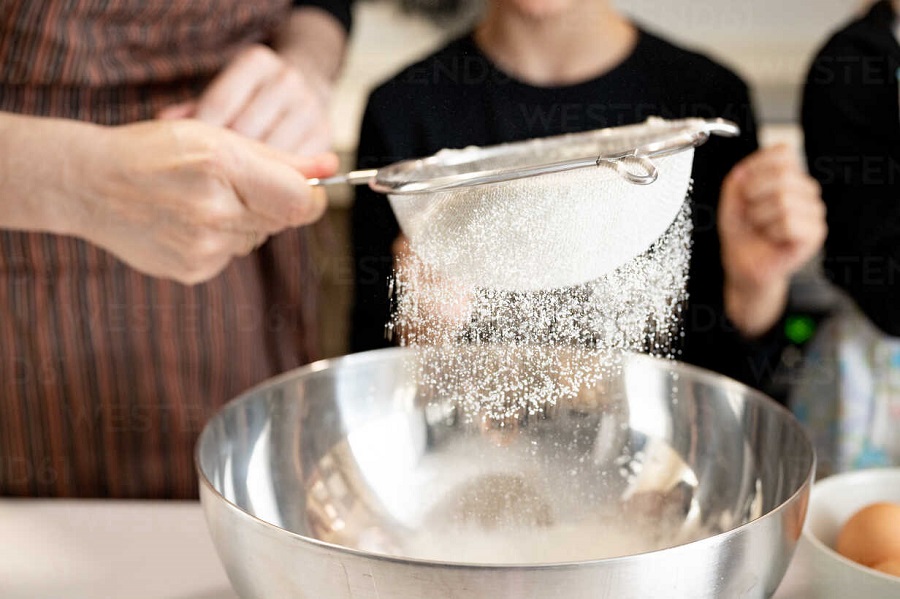 Unrecognizable woman sifting flour into metal bowl while preparing pastry with kids in kitchen at home