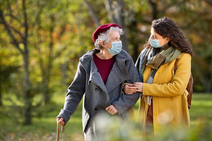 Lovely granddaughter walking with senior woman holding stick in park and wearing mask for safety against covid-19. Happy old grandmother enjoying walking in park with girl. Smiling elderly woman with happy caregiver in park relaxing after quarantine d